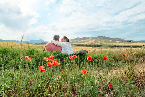 Middle-aged couple sitting hugged in poppies meadow and admir golden wheat field in hot summer sun and blue sky with white clouds with mountains hill landscape in background. photo