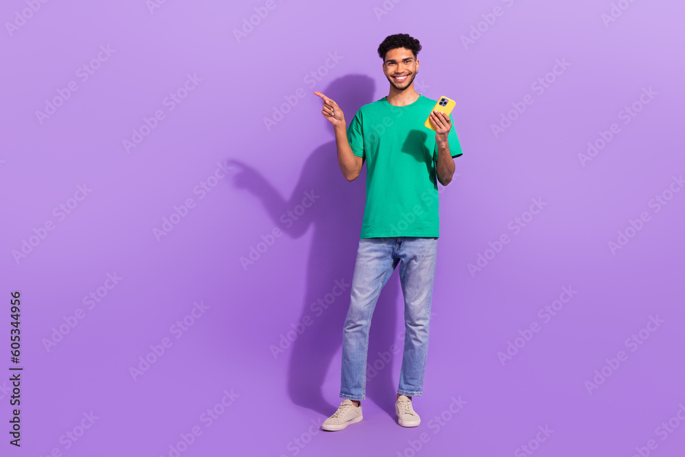 Full body photo of young man blog wear trendy clothes pointing finger empty space phone website promo isolated on purple color background Full body photo of young man blog wear trendy clothes pointing finger empty space phone website promo isolated on purple color background