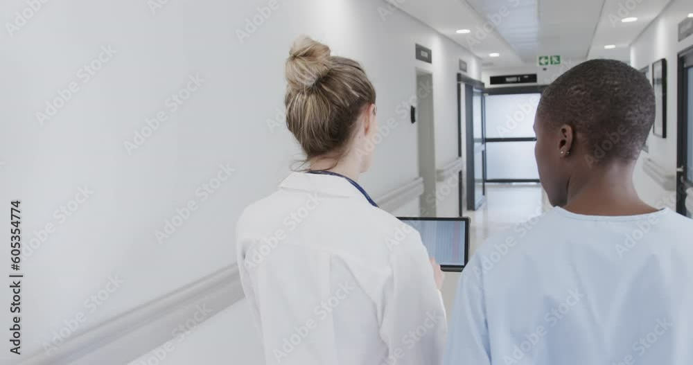Diverse female doctor and nurse in hospital corridor, using tablet and talking, in slow motion