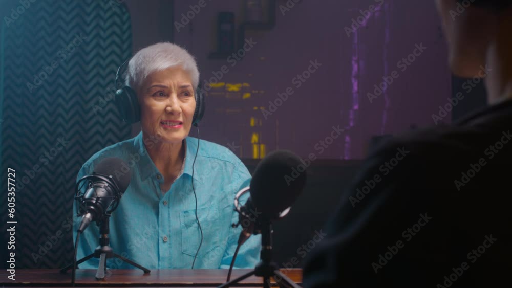 Serious old mature blogger woman in wireless headphones recordings podcast in a professional recording studio. An elderly lady on a radio interview. DJ creates live streaming on the Internet Serious old mature blogger woman in wireless headphones recordings podcast in a professional recording studio. An elderly lady on a radio interview. DJ creates live streaming on the Internet
