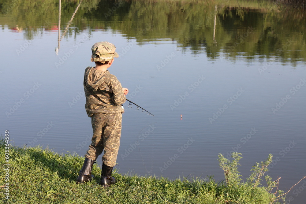 A boy in camouflage clothing and black boots is fishing with a fishing rod on the bank of a quiet river on a spring evening A boy in camouflage clothing and black boots is fishing with a fishing rod on the bank of a quiet river on a spring evening