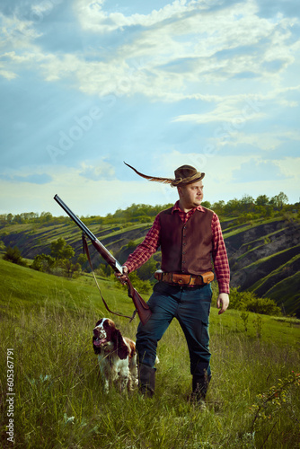 Portrait with man, vintage hunter wearing retro clothes with hat with feather holding gun and standing with dog English springer spaniel over nature landscape background