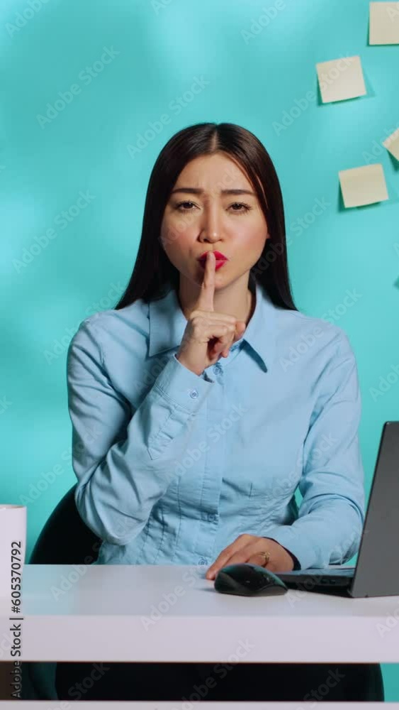 Stern serious businesswoman shushing at camera, holding quiet gesture finger to lips. Strict pensive office employee at bright colourful modern workplace over blue studio background Stern serious businesswoman shushing at camera, holding quiet gesture finger to lips. Strict pensive office employee at bright colourful modern workplace over blue studio background