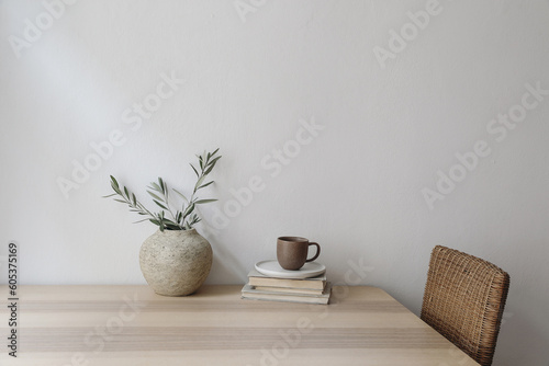 Neutral Mediterranean home design. Vintage vase with olive tree branches, cup of coffee. Books on wooden table, rattan chair. Living room still life. Empty wall copy space. Modern interior, no people.