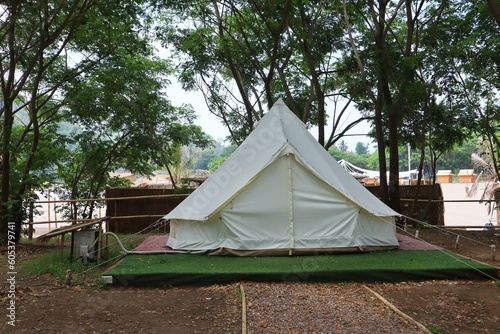 Exterior of white teepee tent at glamping place surrounded by green trees. © Kanomaoi