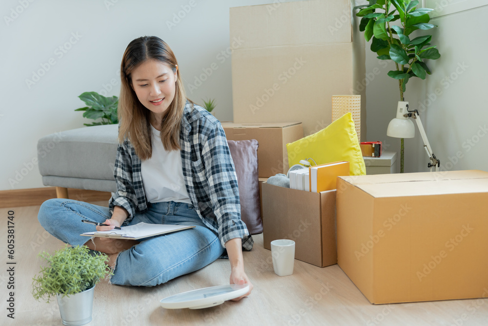 Move house, relocation. Woman using household checklist for new apartment Inside the room are cardboard boxes containing personal belongings and furniture. Move into a house or condominium Move house, relocation. Woman using household checklist for new apartment Inside the room are cardboard boxes containing personal belongings and furniture. Move into a house or condominium