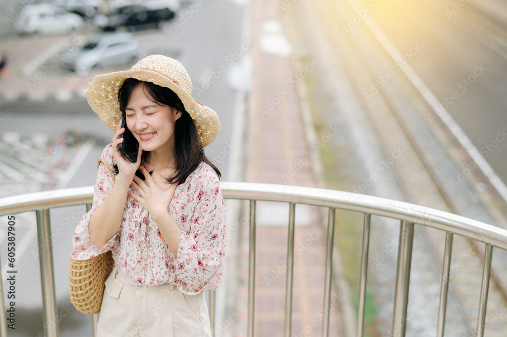 Fototapeta premium young asian woman traveler with weaving basket using mobile phone and standing on overpass with railway background. Journey trip lifestyle, world travel explorer or Asia summer tourism concept.
