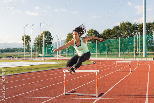 Young overweight woman running and jumping over hurdles on an athletics track on a sunny summer day