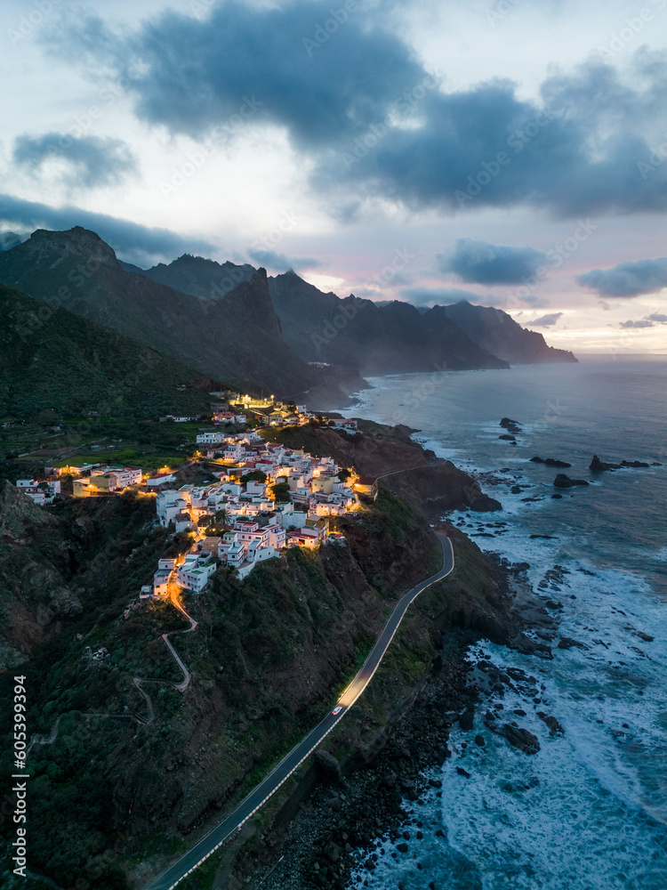 Drone shot of a little white village on the ocean with orange lights at the blue hour surrounded by mountains in northern Tenerife