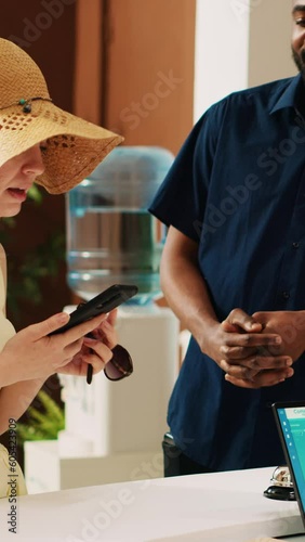 Vertical video Diverse couple using mobile nfc payment with smartphone next to pos terminal, paying for room reservation at tropical summer resort. Tourists doign check in at front desk reception. photo