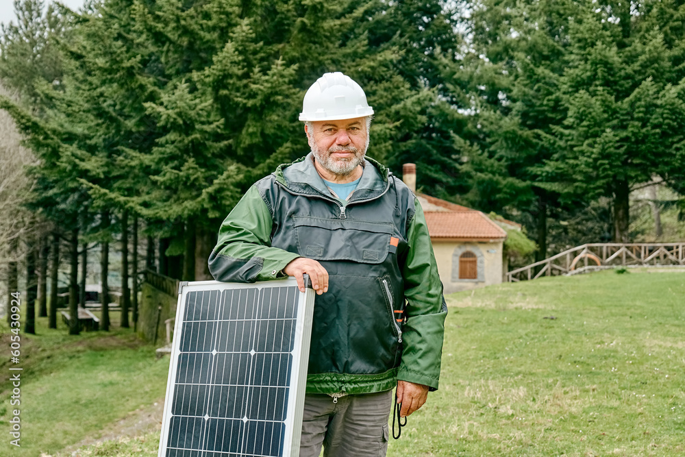 Middle aged bearded technician with voltaic solar panel near house in rural zone. Stand-alone photovoltaic solar panel system. Renewable ecological cheap green energy production. Energy independence. Middle aged bearded technician with voltaic solar panel near house in rural zone. Stand-alone photovoltaic solar panel system. Renewable ecological cheap green energy production. Energy independence.