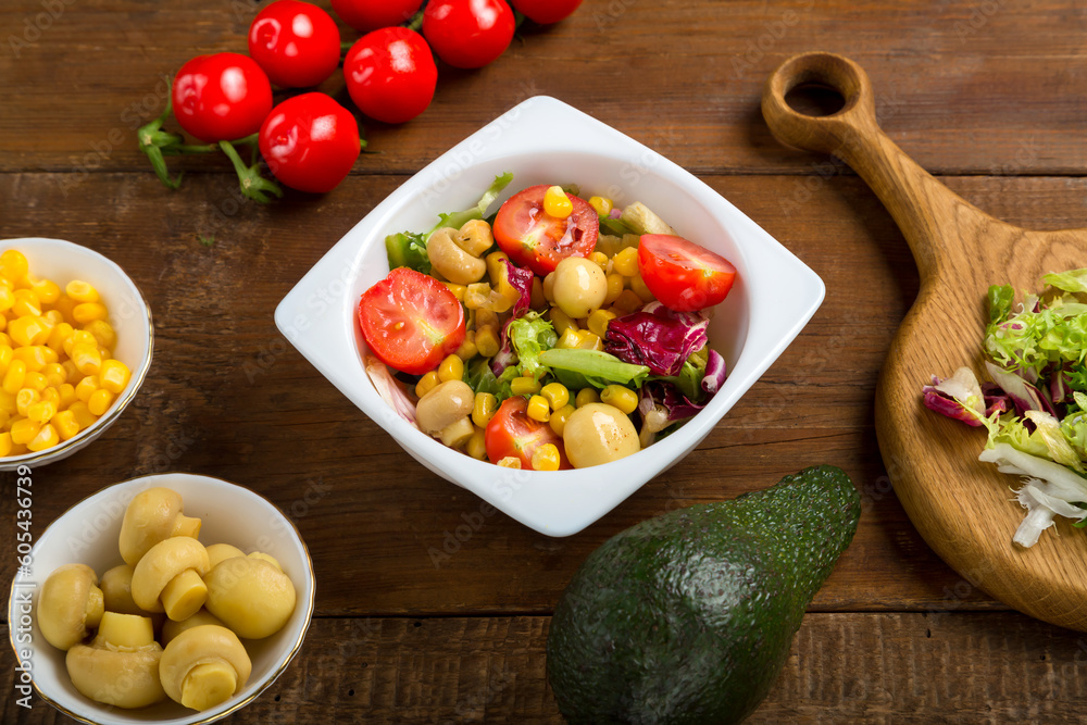Vegetable salad with champignon corn and cherry tomato with avocado and olive oil in a white plate on a wooden table next to all the ingredients. Vegetable salad with champignon corn and cherry tomato with avocado and olive oil in a white plate on a wooden table next to all the ingredients.