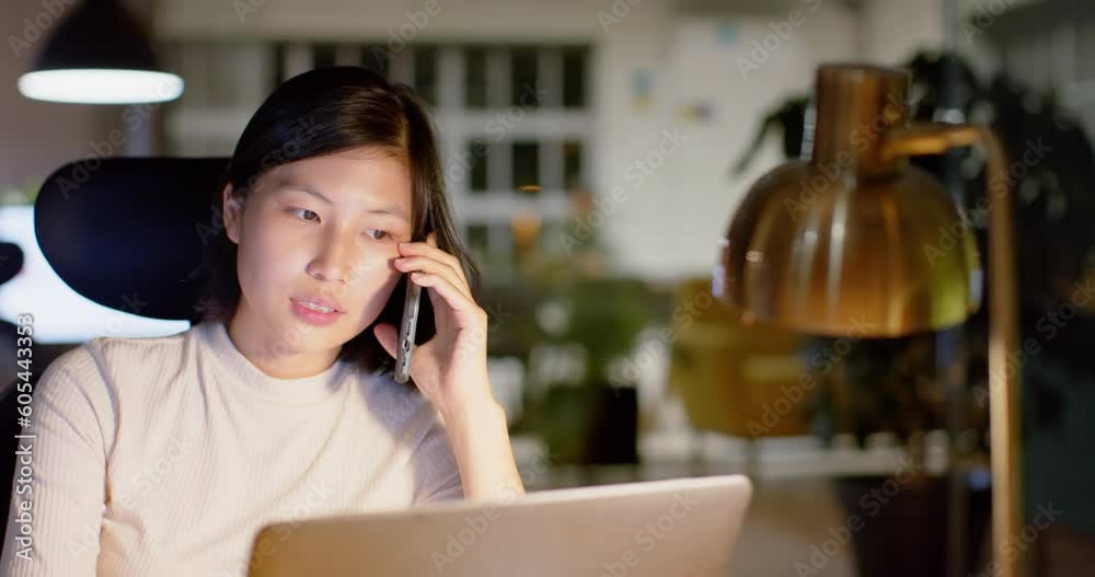 Asian casual businesswoman talking on smartphone and using laptop in office at night, slow motion