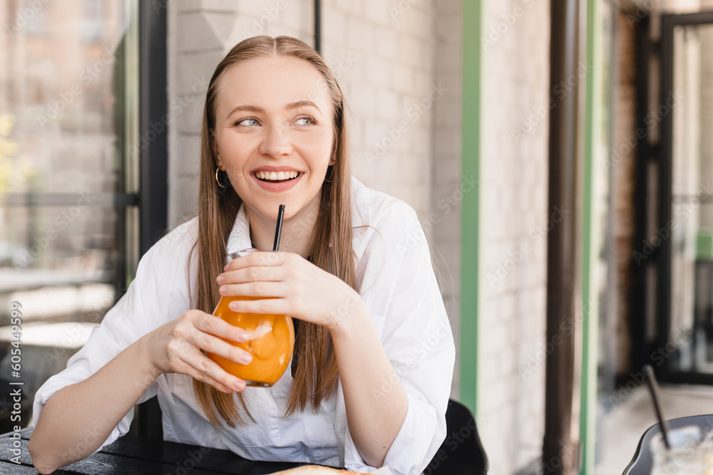 Close-up of young caucasian woman enjoying drinking orange lemonade sitting in cafe. Blonde in white shirt quenches her thirst summer. Relaxation concept. Happy laughing woman. Close-up of young caucasian woman enjoying drinking orange lemonade sitting in cafe. Blonde in white shirt quenches her thirst summer. Relaxation concept. Happy laughing woman.