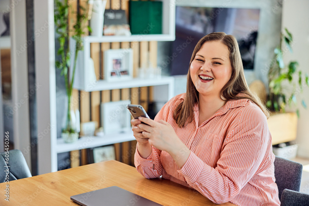 A portrait of a laughing curvy woman using a mobile phone and sitting in front of a laptop.