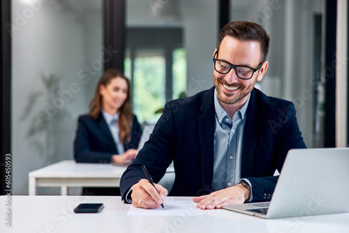 Smiling man dressed elegantly, working at the office, female colleague sitting in the background.