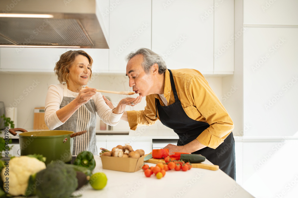 Cooking together concept. Senior woman feeding her husband while preparing dinner in kitchen, making delicious meal