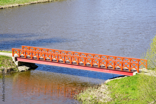 A small orange pedestrian bridge over a river with a steel girder base and wooden railings
