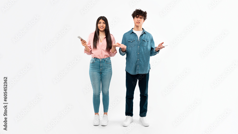 Obraz premium Displeased Young Couple Using Mobile Phones Standing Over White Background