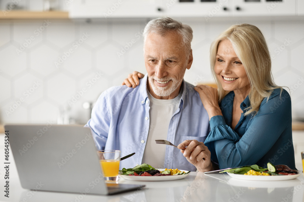 Smiling Elderly Spouses Relaxing With Laptop While Having Lunch In Kitchen