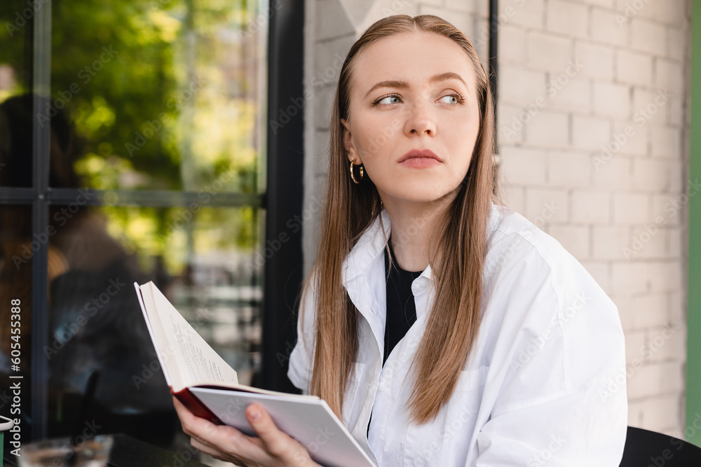 Pensive woman in casual outfit resting on chair at table with cup reading interesting book, chilling in restaurant. Blonde calm woman studying in cafe, read novel, historical book. Think about it. Pensive woman in casual outfit resting on chair at table with cup reading interesting book, chilling in restaurant. Blonde calm woman studying in cafe, read novel, historical book. Think about it.