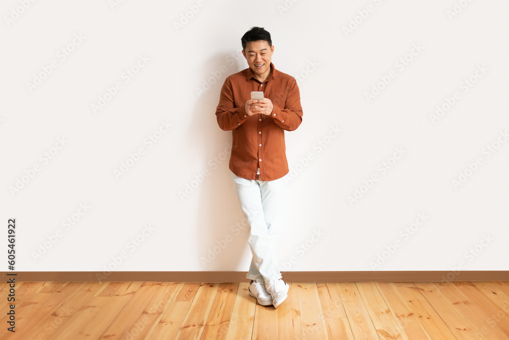 Mobile lesiure concept. Asian middle aged man using cellphone, surfing internet online standing against white wall Mobile lesiure concept. Asian middle aged man using cellphone, surfing internet online standing against white wall