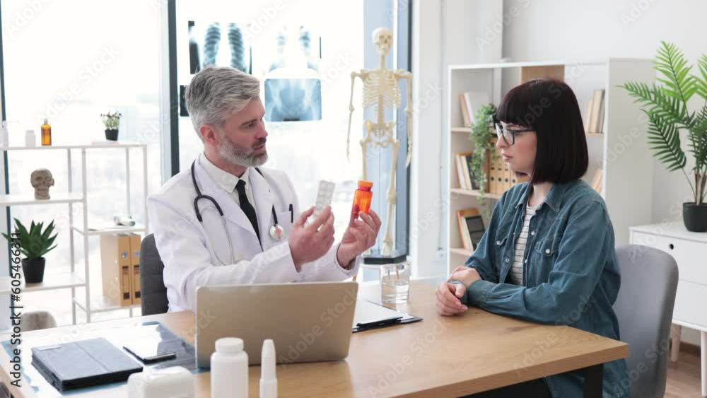 Mature male in white coat holding medications while talking to beautiful woman in casual wear sitting at office desk. Efficient therapist prescribing pills for easing symptoms of neck pain in clinic. Mature male in white coat holding medications while talking to beautiful woman in casual wear sitting at office desk. Efficient therapist prescribing pills for easing symptoms of neck pain in clinic.