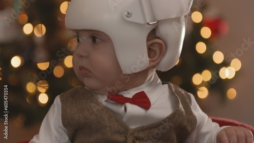 8 Month Old Baby Wearing Therapy Helmet Christmas Pose Tilt Up Look Left. slow motion christmas pose of an 8 month old sitting in front of christmas tree wearing a cranial therapy helmet tilt up photo