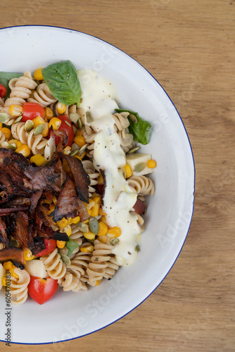 Top view of a Fusilli pasta salad with basil, crispy bacon, corn, cheese, tomato and a poached egg, in a white bowl on the wooden restaurant table.
