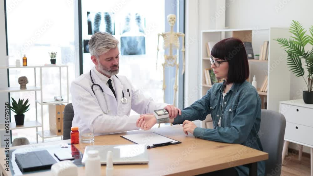 Mature caucasian man in lab coat putting electronic device on woman's wrist during check-up in consulting room. General practitioner applying pulse oximeter for female patient at health clinic. Mature caucasian man in lab coat putting electronic device on woman's wrist during check-up in consulting room. General practitioner applying pulse oximeter for female patient at health clinic.
