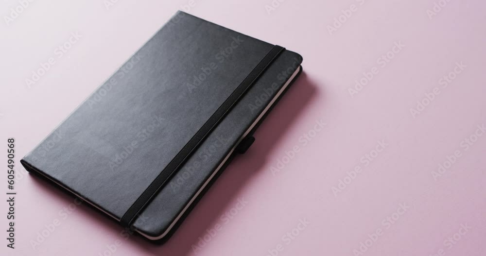 Close up of closed black notebook with copy space on pink background in slow motion Close up of closed black notebook with copy space on pink background in slow motion