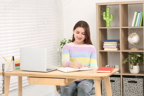 Cute girl writing in notepad near laptop at desk in room. Home workplace