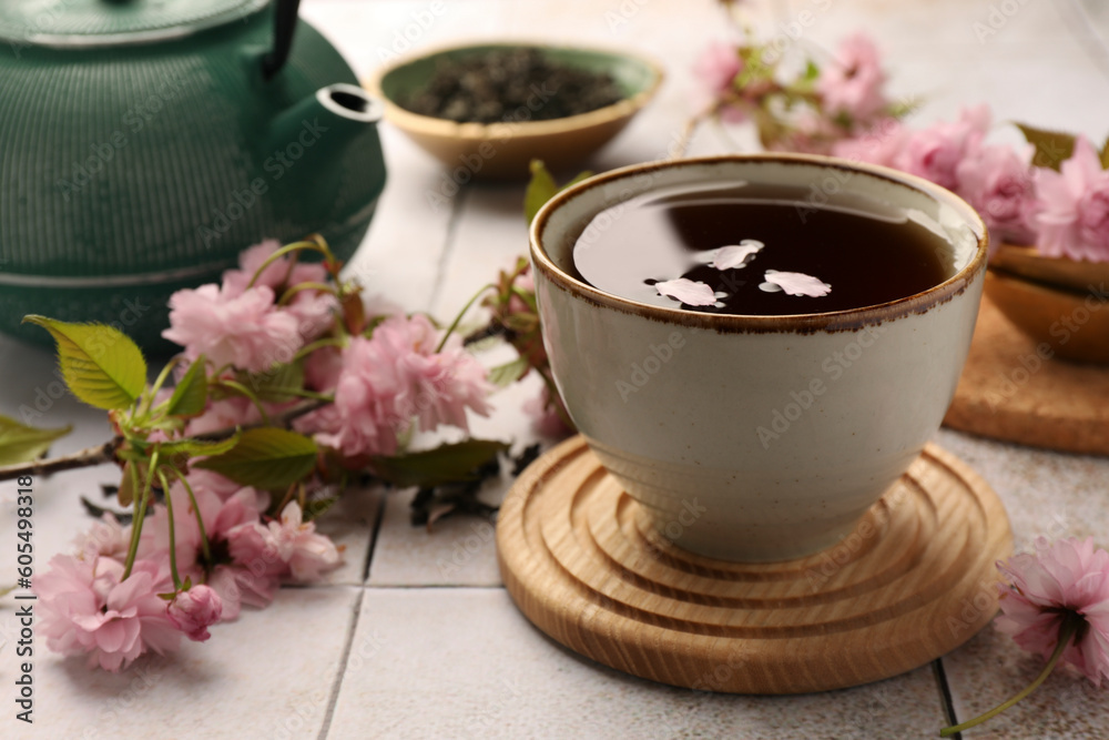 Fototapeta premium Traditional ceremony. Sakura petals in cup of brewed tea, teapot and flowers on tiled table, closeup