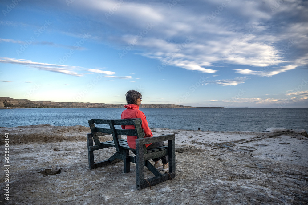 Senior woman contemplates the sea sitting on a bench on the rocks on a partly cloudy day, in Puerto Pirámides, Chubut, Argentina.
