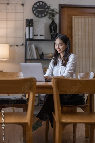 Business And Education Concept. Smiling young asian woman sitting at desk working on laptop writing letter in paper documents, free copy space. Happy millennial female studying using laptop © David
