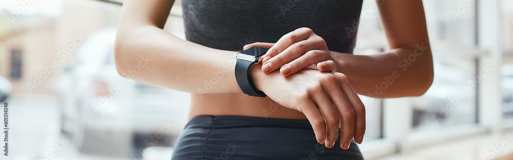 Checking all updates. Cropped photo of beautiful young woman looking at her sport bracelet while standing in front of window at gym