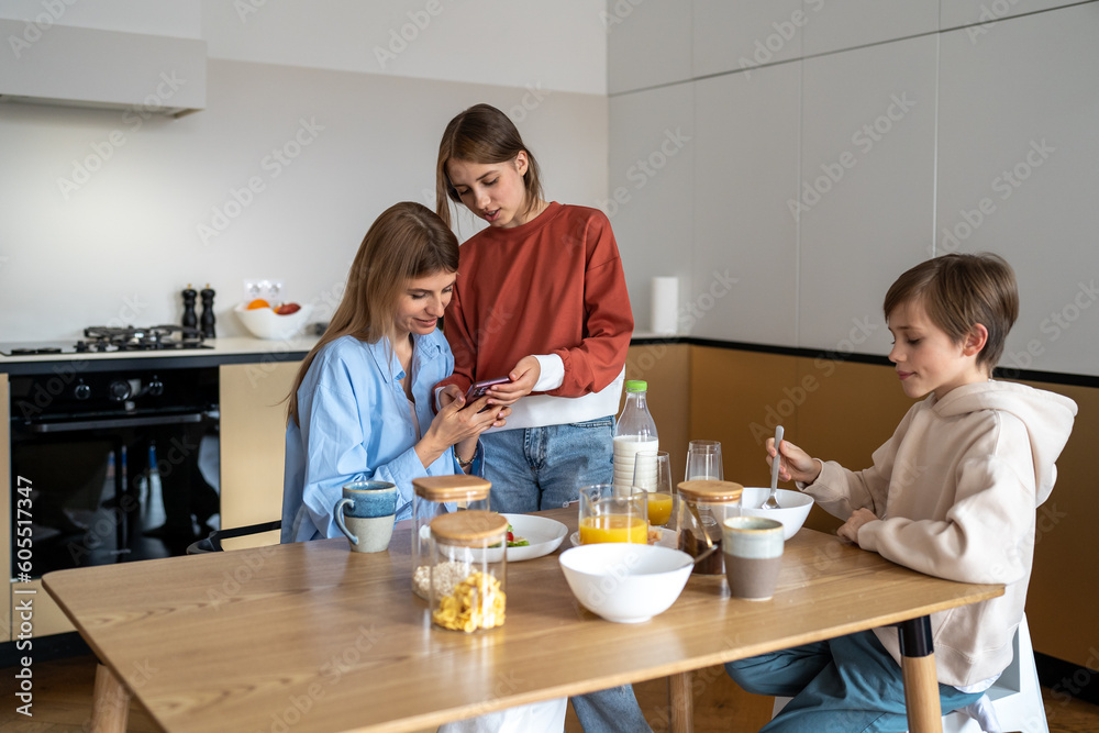 Teen daughter showing new video for social media on smartphone to mom during family breakfast on kitchen. Mother son eating sitting at table. Motherhood, parenting, rising children, mutual support. Teen daughter showing new video for social media on smartphone to mom during family breakfast on kitchen. Mother son eating sitting at table. Motherhood, parenting, rising children, mutual support.
