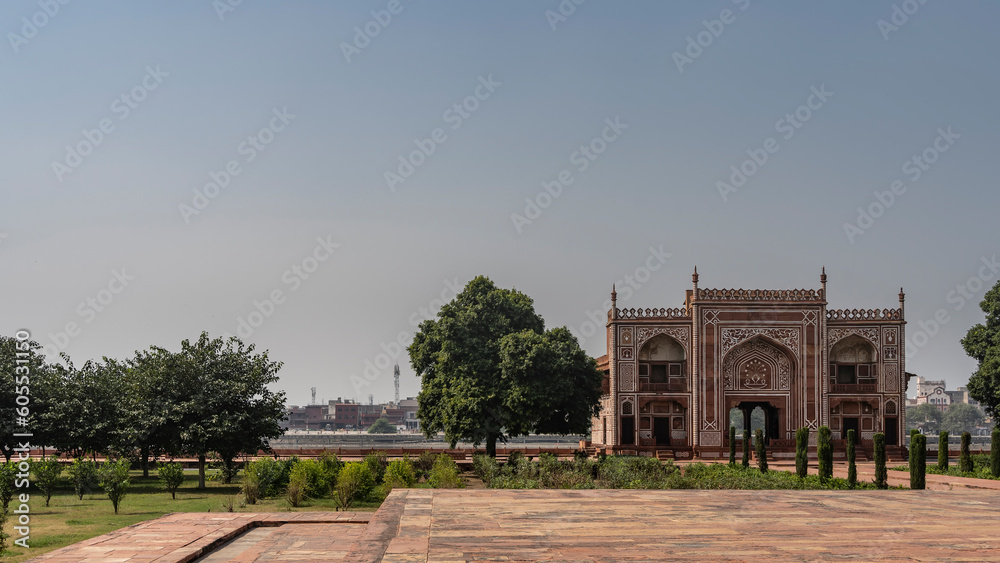 The ancient building in the complex tomb of Itmad-Ud-Daulah built of red sandstone, decorated with white ornaments. Arches, balconies, spires are visible. Green plants in the garden.  India. Agra