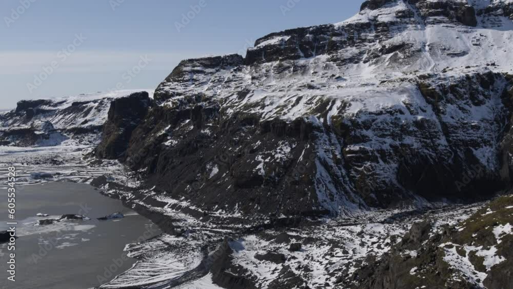 Drone clip of snowy steep rocky cliffs next to icy lake in Iceland on sunny day Drone clip of snowy steep rocky cliffs next to icy lake in Iceland on sunny day