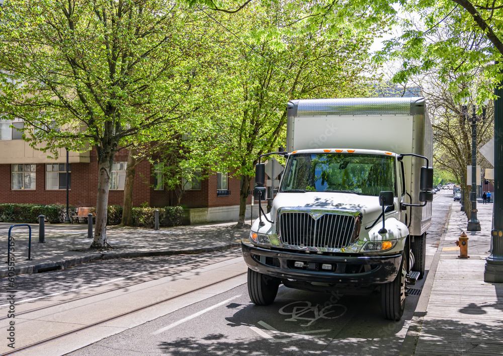 Obraz premium In demand white middle duty rig semi truck with day cab and box trailer for local delivery standing on the urban city street with trees shadow