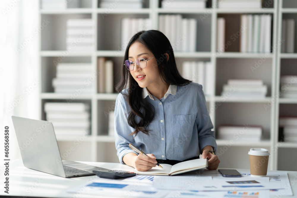 Modern working lifestyle Asian businesswoman sits at desk with laptop online using internet and notepad happily recording receipt contact information online delivery orders.