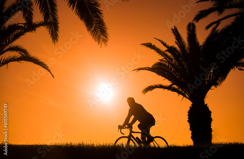 male riding without hands. alone black Silhouette of man cycling along horizon line. one Silhouette palm trees on the hill. orange sun set sky at yellow sunset. sun rays. grass meadow. rent cross bike