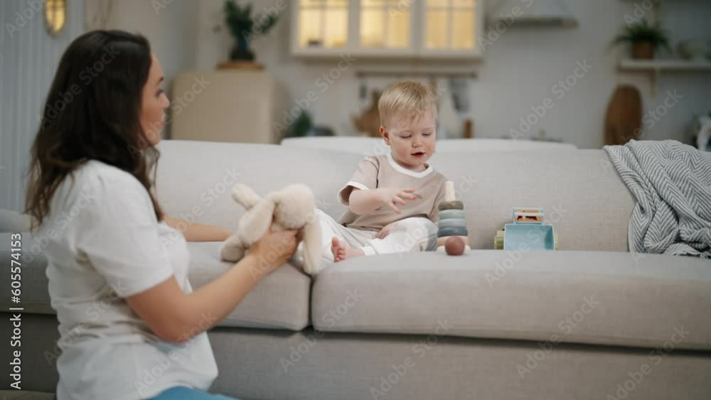 Playful positive mother playing teddy hare and wooden pyramid with baby boy toddler sitting on couch at home. Subject-manipulative games for kids development and education. Motherhood, babycare. Playful positive mother playing teddy hare and wooden pyramid with baby boy toddler sitting on couch at home. Subject-manipulative games for kids development and education. Motherhood, babycare.