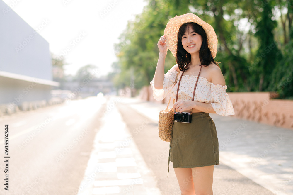 Portrait of asian young woman traveler with weaving hat and basket and a camera standing by the street. Journey trip lifestyle, world travel explorer or Asia summer tourism concept.