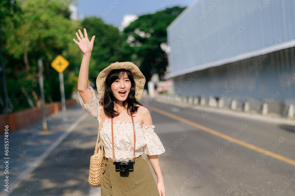 Portrait of asian young woman traveler with weaving hat and basket and a camera waving hand to friend by the street. Journey trip lifestyle, world travel explorer or Asia summer tourism concept.