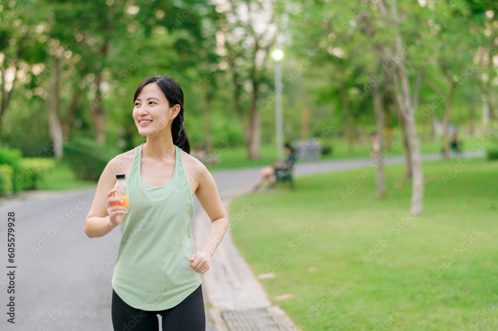 Female jogger. Fit young Asian woman with green sportswear drinking organic orange juice after running and enjoying a healthy outdoor. Fitness runner girl in public park. Wellness being concept Female jogger. Fit young Asian woman with green sportswear drinking organic orange juice after running and enjoying a healthy outdoor. Fitness runner girl in public park. Wellness being concept