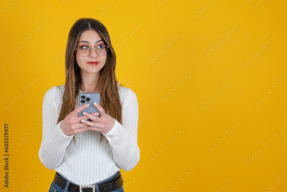 Using smartphone, portrait of beautiful caucasian woman using smartphone. Standing over yellow studio background. Smiling and looking copy space area without turning her head. Ad concept idea. Using smartphone, portrait of beautiful caucasian woman using smartphone. Standing over yellow studio background. Smiling and looking copy space area without turning her head. Ad concept idea.