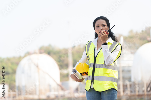female engineer with hardhat with petrochemical factory background. asian woman holding tablet, plan and Walkie Talkie.