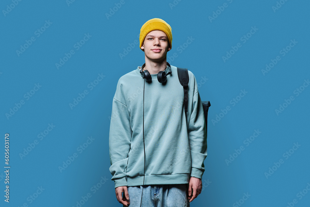 Young male in yellow cap sweatshirt with headphones backpack on blue background Young male in yellow cap sweatshirt with headphones backpack on blue background
