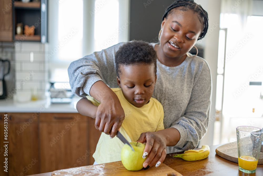Fototapeta premium Cheerful African-American mother and son in the kitchen preparing a healthy apple snack